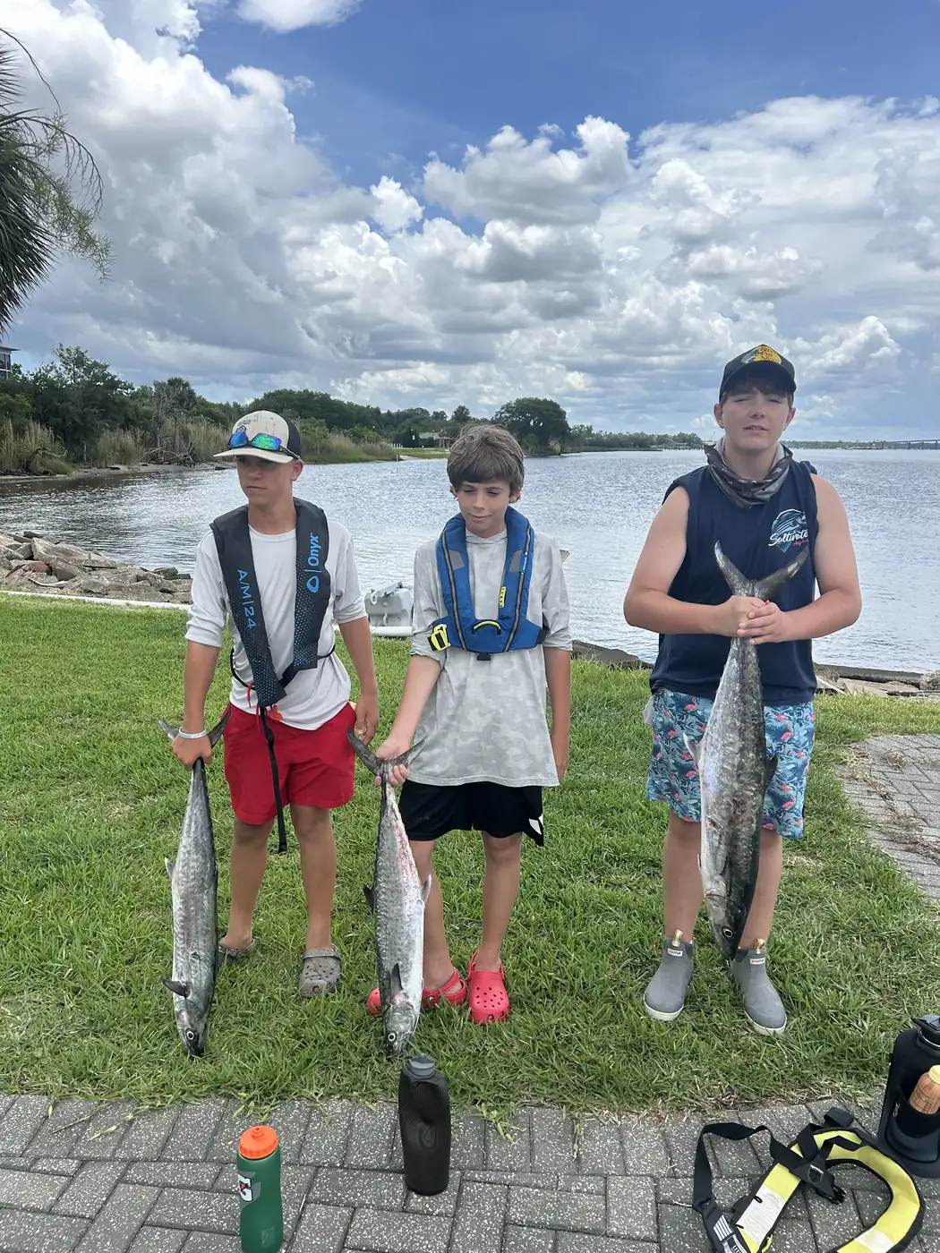 Three campers display their fish together on grass beside the waterfront.