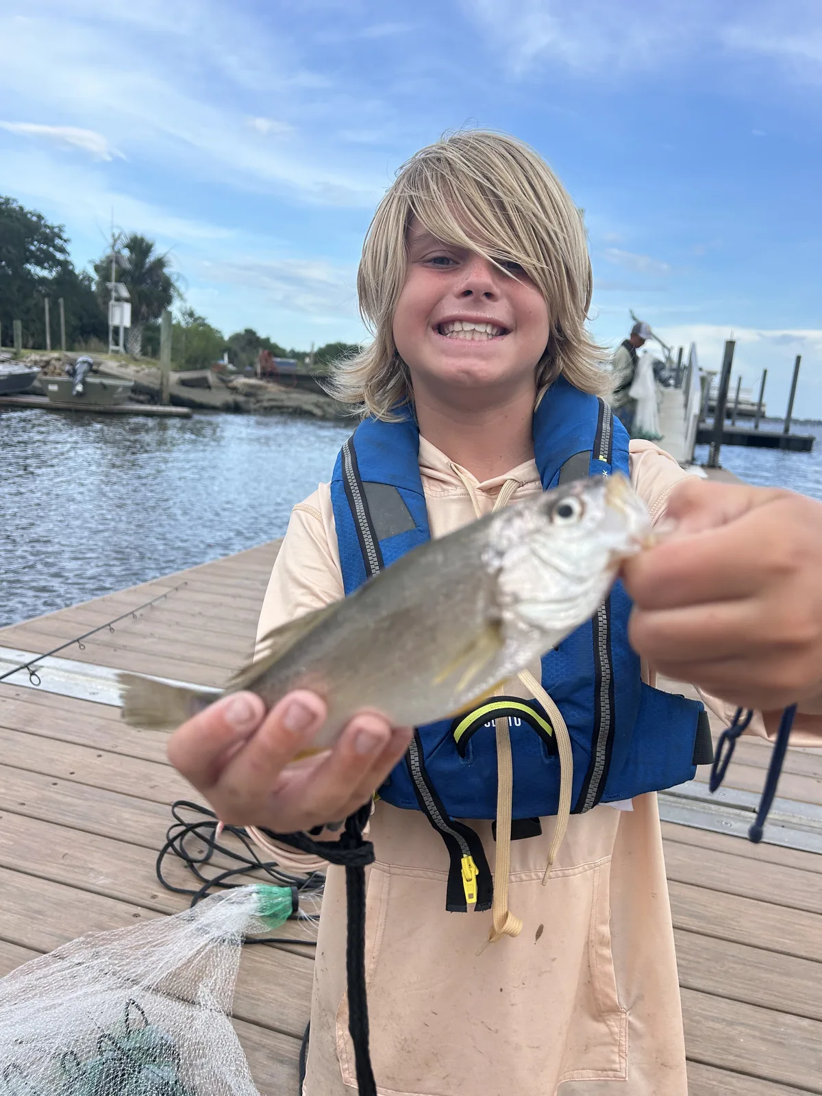 A smiling camper holds a small fish while standing on a dock.