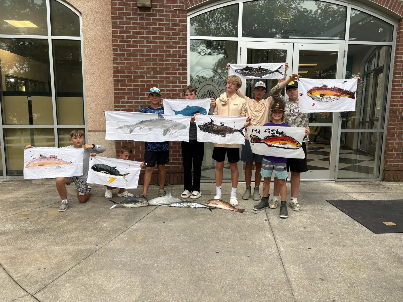 A group of campers holds colorful fish-print artwork outside a building.