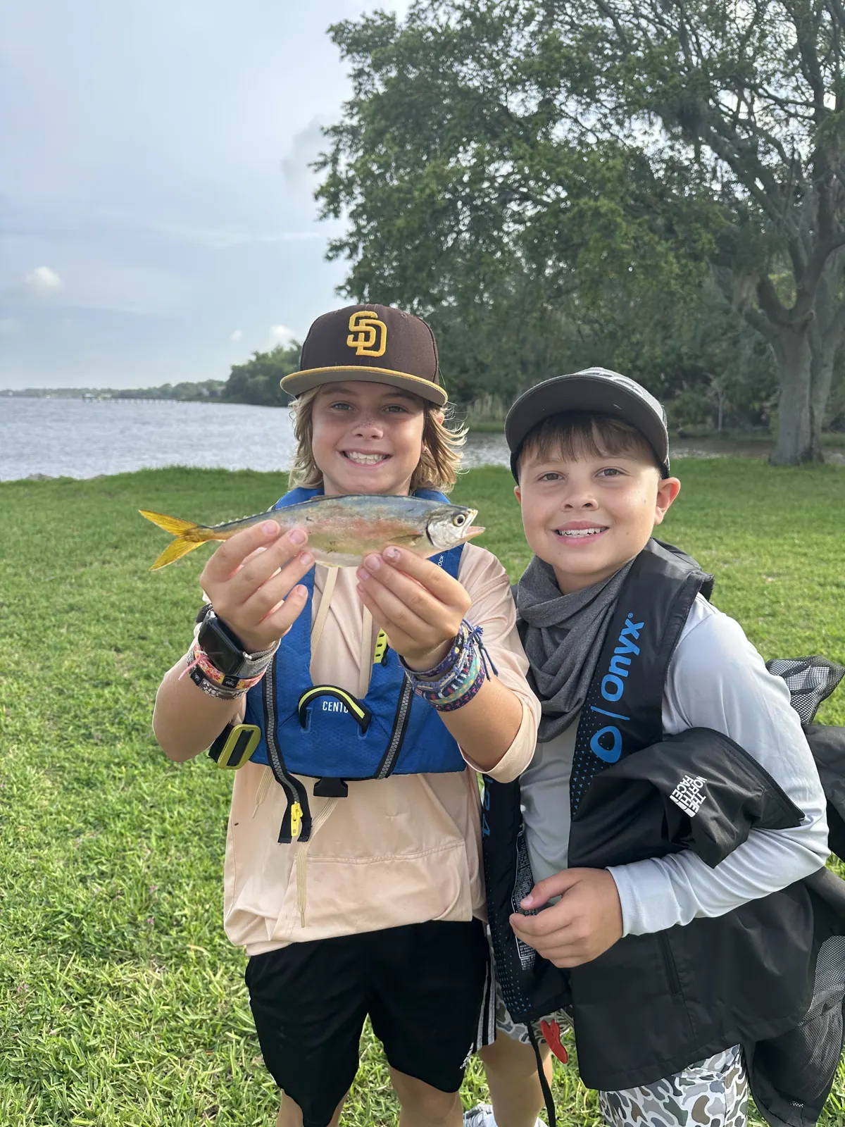 Two campers smile while one holds a small fish beside the waterfront.