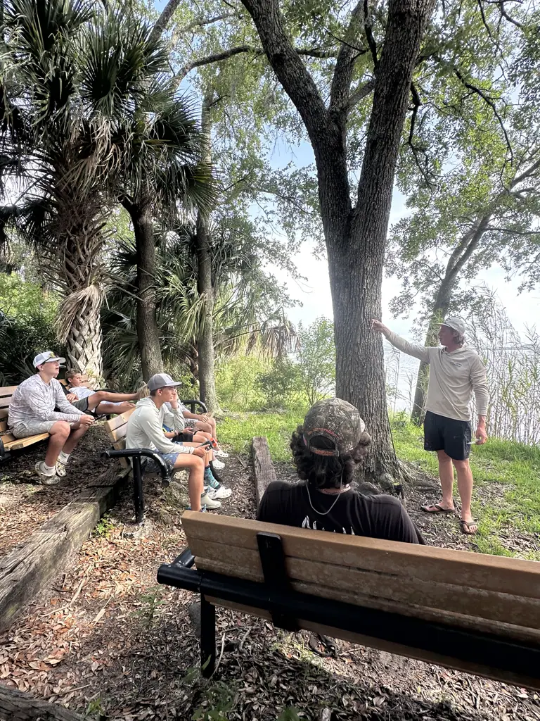 Campers sit on benches while an adult stands beside a tree near the water.